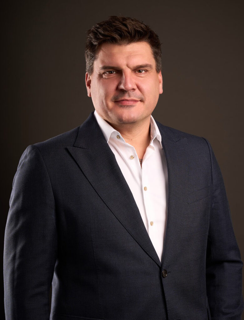 Portrait of the contact expert Slobodan Bozinovski in a dark blazer and white shirt, standing against a neutral background, looking directly at the camera.