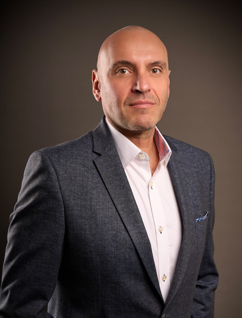 Portrait of Daniel Atanasovski in a dark blazer and white shirt, standing against a neutral background, looking directly at the camera.